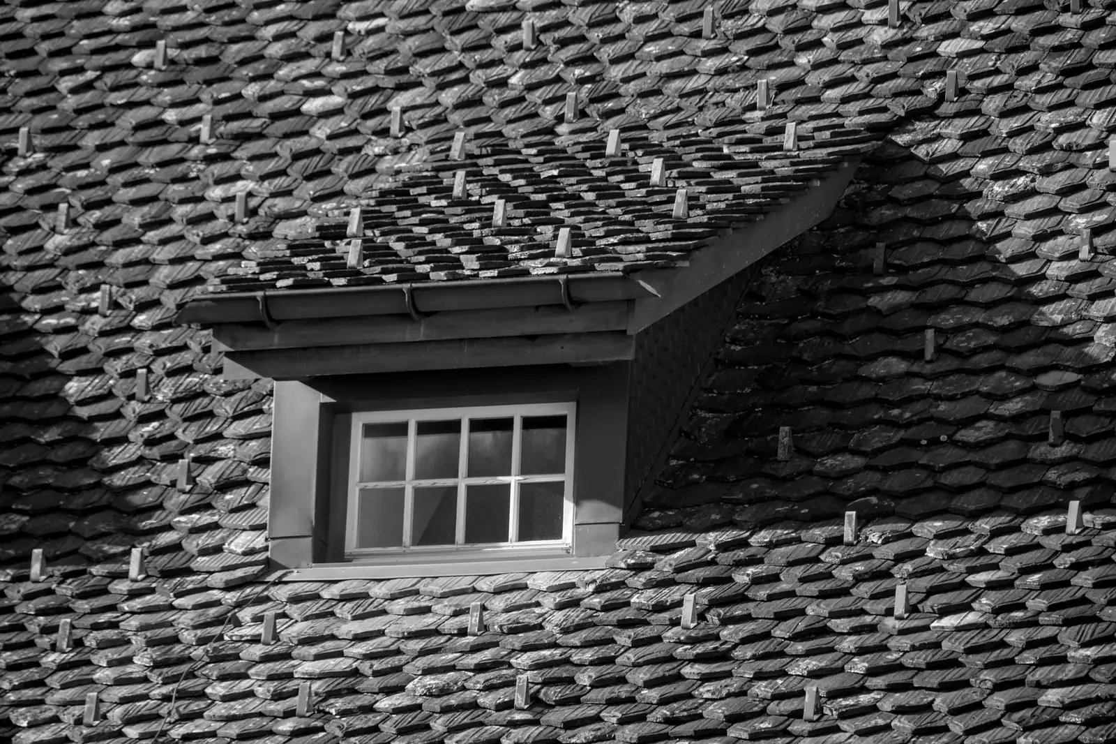 Lucarne dont le petit toit est presque horizontal, posée sur un versant d'ardoises, photographiée en noir et blanc